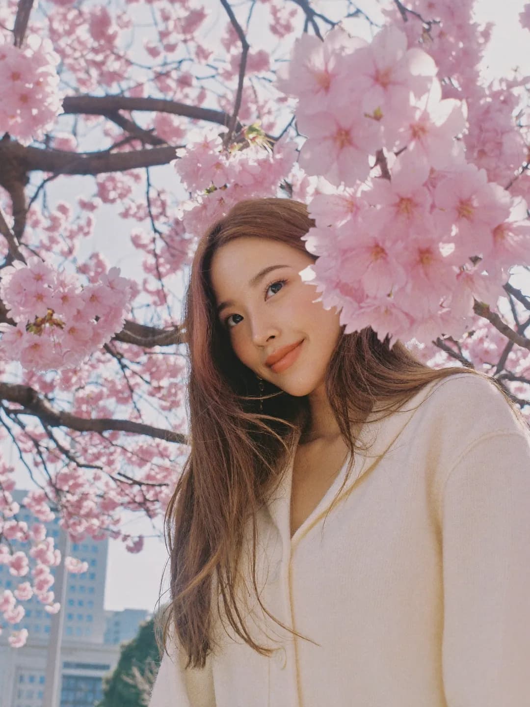 Seorang wanita berambut panjang tersenyum di bawah bunga sakura berwarna merah jambu pucat.