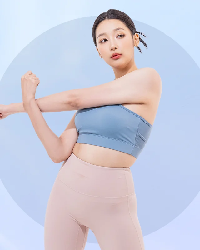 Seorang wanita muda memakai atasan sukan biru dan legging bej, meregangkan sebelah lengan merentasi dada, berlatarkan warna biru pucat.