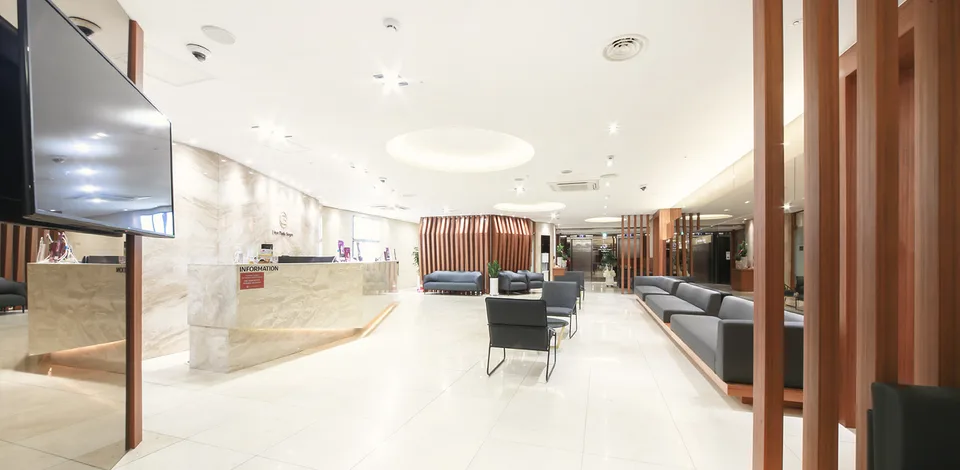 Quầy thông tin và khu vực chờ tại Urban Plastic Surgery, phòng khám đối tác của chúng tôi ở Seoul, với sofa màu xám, tường ốp lam gỗ và một màn hình treo trên tường.